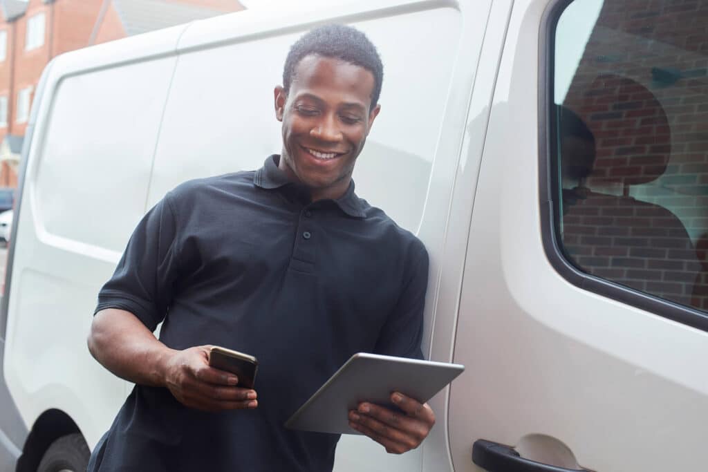 A young courier stands next to his van while using a digital tablet and smart phone to update delivery status.