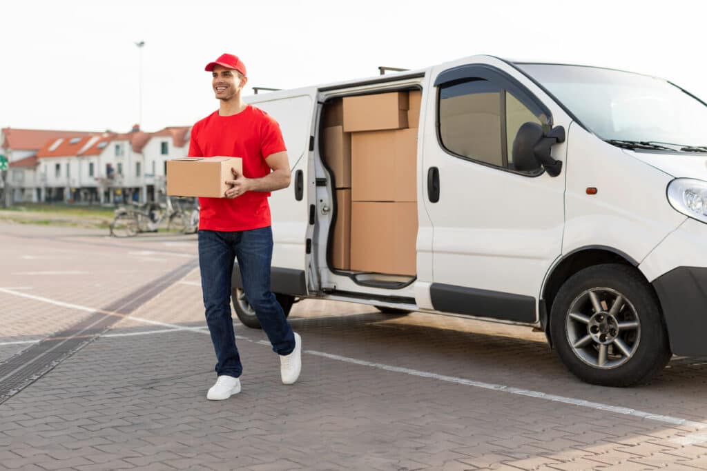 A last-mile courier in a red t-shirt and hat arrives at his destination and carries a delivery to the customer’s door.